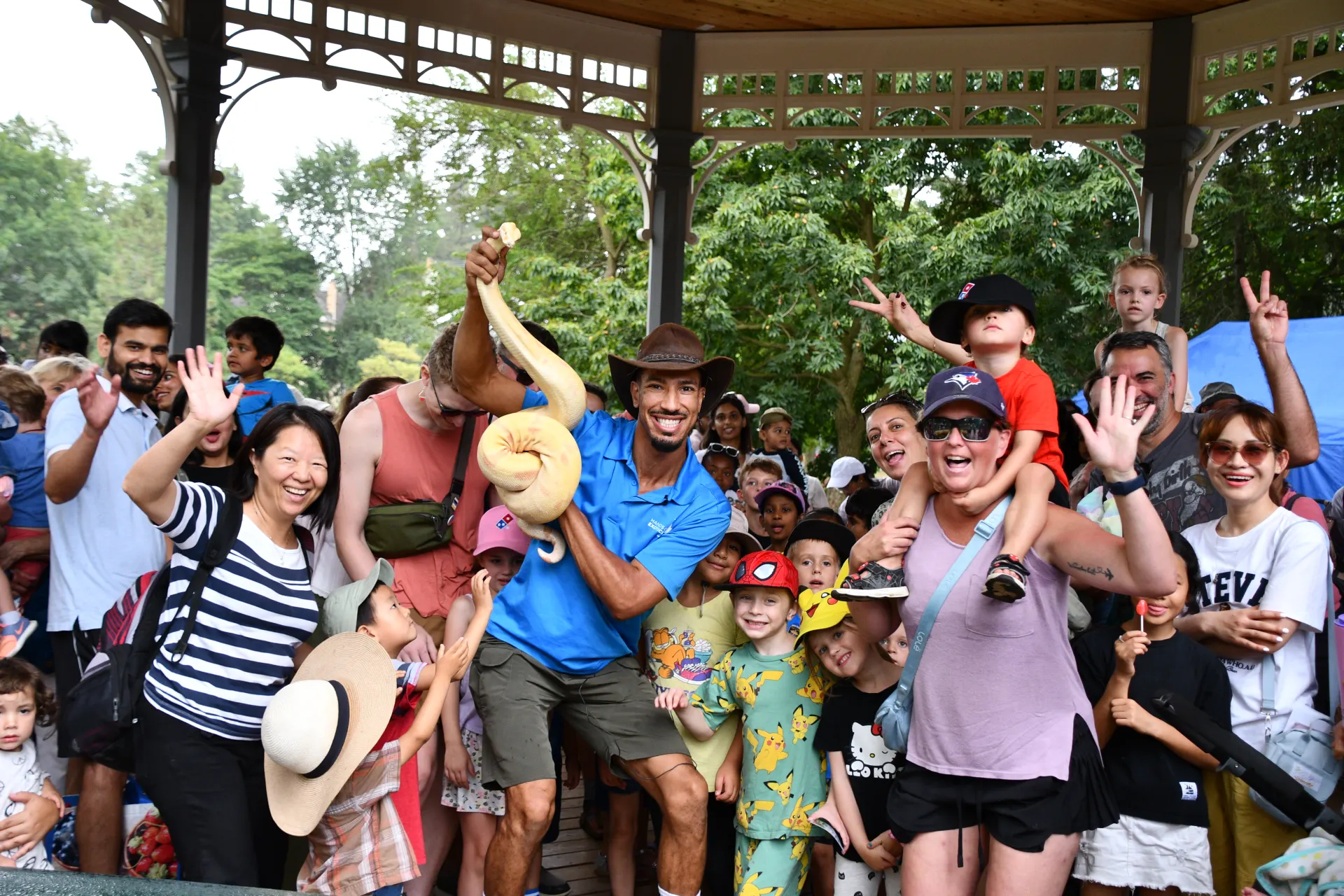 Seth Falk in a cyan polo confidently holding up a large albino python under a wooden pavilion, surrounded by a smiling crowd of families and excited children at a community event