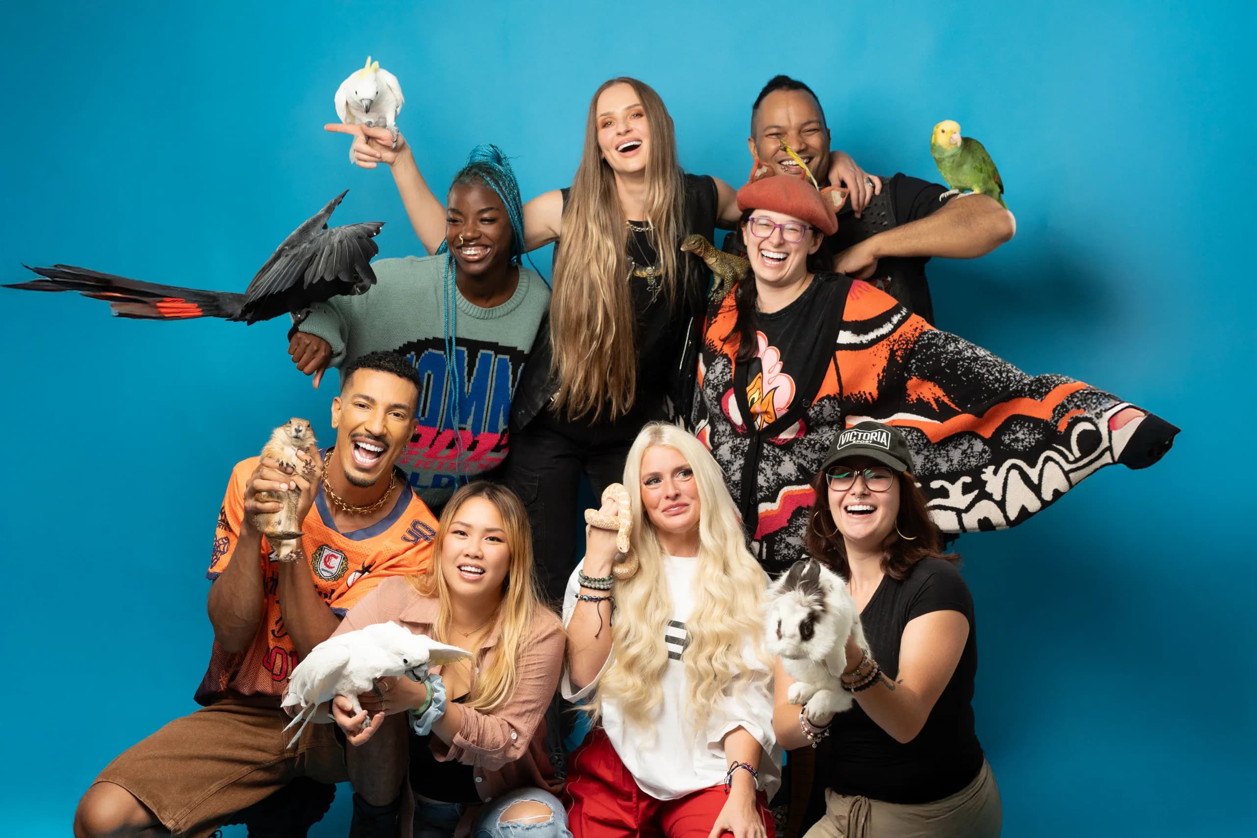 The Hands On Exotics team — eight handlers posing with cockatoos, a raven, a parrot, a lizard, a prairie dog, a python, and a rabbit against a bright cyan backdrop.